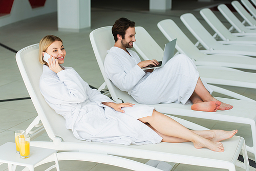 Cheerful woman talking on smartphone near orange juice and boyfriend with laptop in spa center