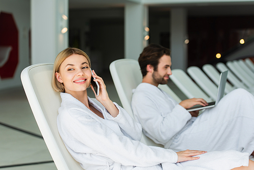 Positive woman in bathrobe talking on smartphone near blurred boyfriend with laptop in spa center