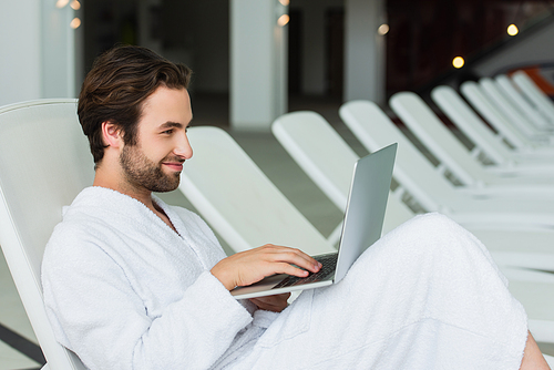 Smiling man in white bathrobe using laptop on deck chair in spa center