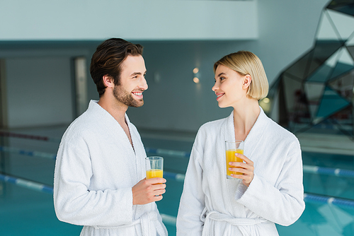 Positive couple in white bathrobes holding orange juice in spa center