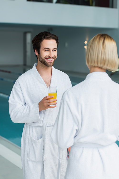 Smiling man in white bathrobe holding glass of orange juice near blurred girlfriend and swimming pool in spa center