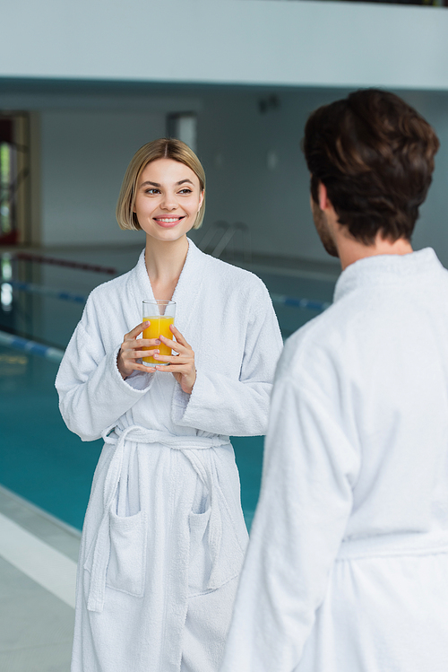 Cheerful woman holding glass of orange juice near blurred boyfriend in white bathrobe in spa center