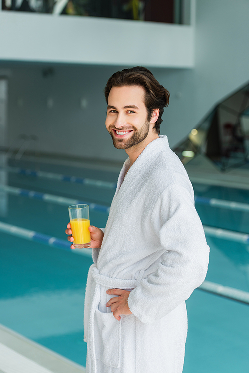 Young man in white bathrobe holding glass of orange juice near blurred swimming pool in spa center