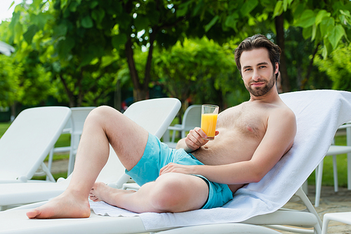 Young man in swimming trunks holding orange juice on deck chair on resort