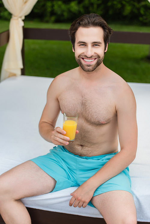 Smiling man in swimming trunks holding orange juice on lounge bed on resort