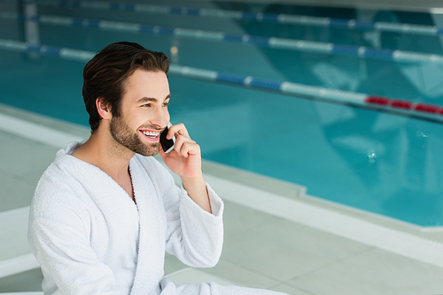 Smiling man in bathrobe talking on mobile phone near blurred swimming pool in spa center