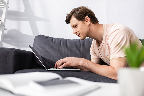 Selective focus of handsome freelancer using laptop on couch at home, earning online concept