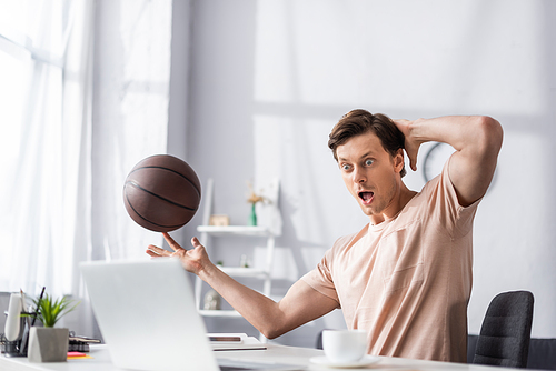 Selective focus of shocked man holding basketball and looking at laptop on table, concept of earning online
