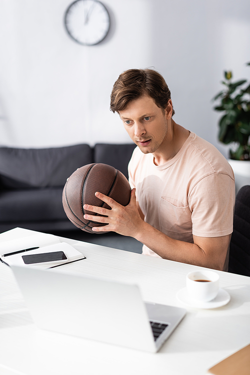 Selective focus of handsome freelancer holding basketball and looking at laptop near cup of coffee on table, earning online concept
