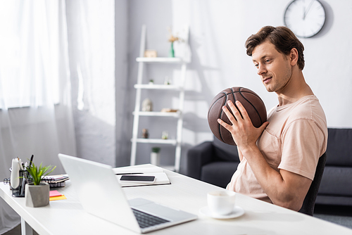 Selective focus of handsome man holding basketball near laptop and stationery on table at home, concept of earning online