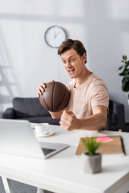 Selective focus of cheerful man holding basketball and showing yeah gesture near laptop on table, earning online concept