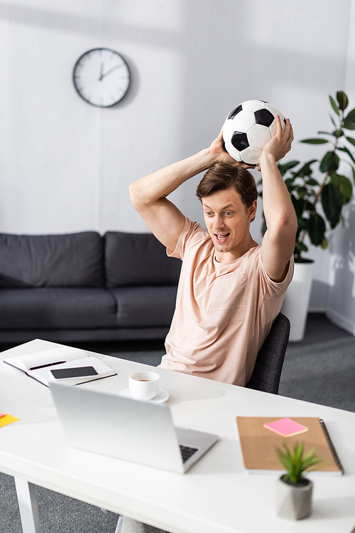 Selective focus of positive man holding football and looking at laptop on table at home, concept of earning online