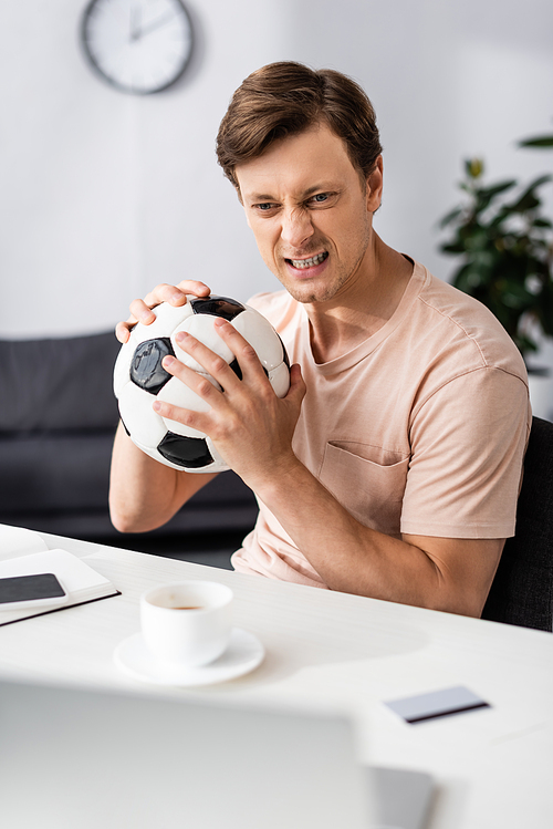 Selective focus of angry man holding football near gadgets and credit card on table, concept of earning online