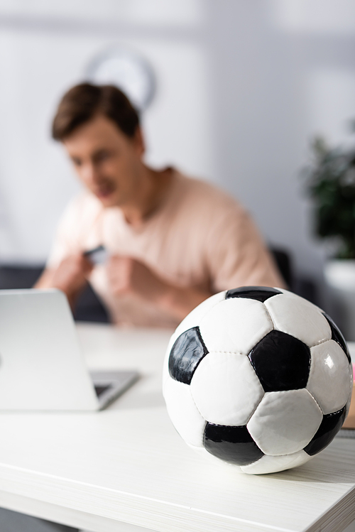 Selective focus of football on table and man sitting near laptop at home, concept of earning online