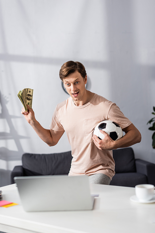 Selective focus of excited man holding cash and football near laptop on table at home, concept of earning online
