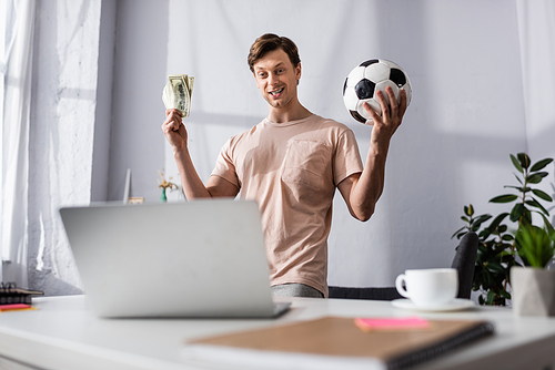 Selective focus of cheerful man holding football and money near laptop at home, concept of earning online