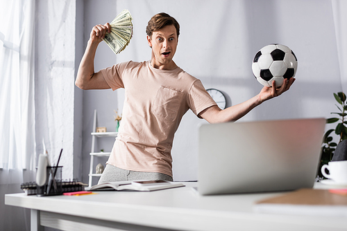 Selective focus of shocked man with cash and football looking at laptop in living room, concept of earning online