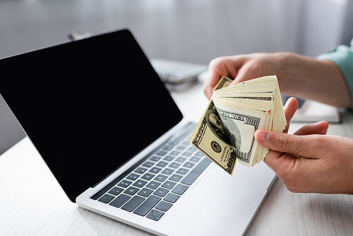 Cropped view of man holding dollar banknotes near laptop with blank screen on table, earning online concept