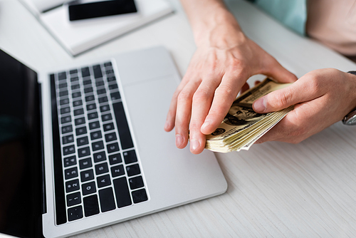 Cropped view of man holding cash near laptop on table, earning online concept