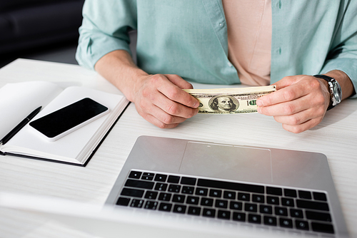 Cropped view of man holding dollars near laptop and smartphone with blank screen on table, concept of earning online