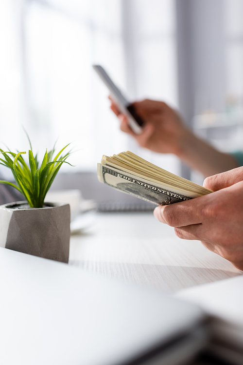 Cropped view of man holding money and using smartphone near plant on table at home, earning online concept
