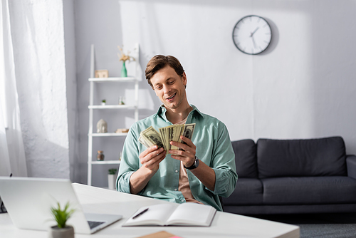 Selective focus of smiling man counting money near laptop and notebook on table in living room, concept of earning online