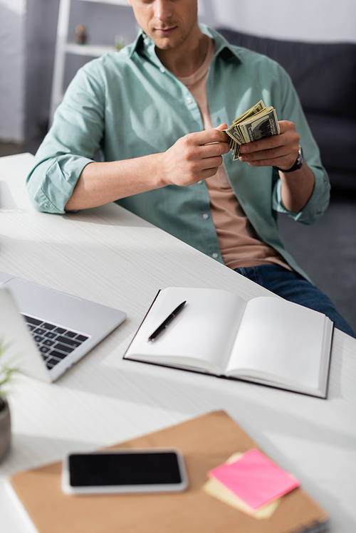 Cropped view of man holding cash near digital devices and notebooks on table at home, earning online concept