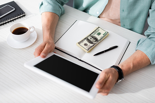 Cropped view of freelancer holding digital tablet near coffee and dollar banknotes on notebook at table, concept of earning online