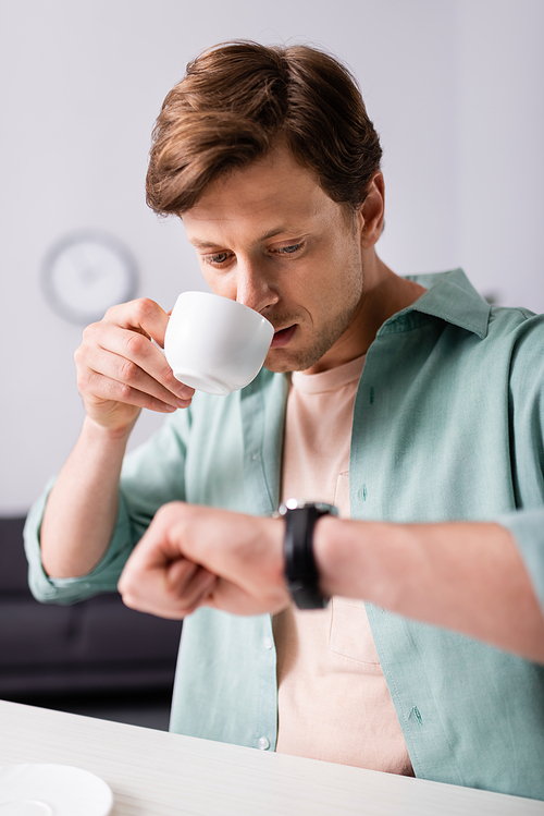 Selective focus of handsome man drinking coffee and checking time on wristwatch at home, concept of time management
