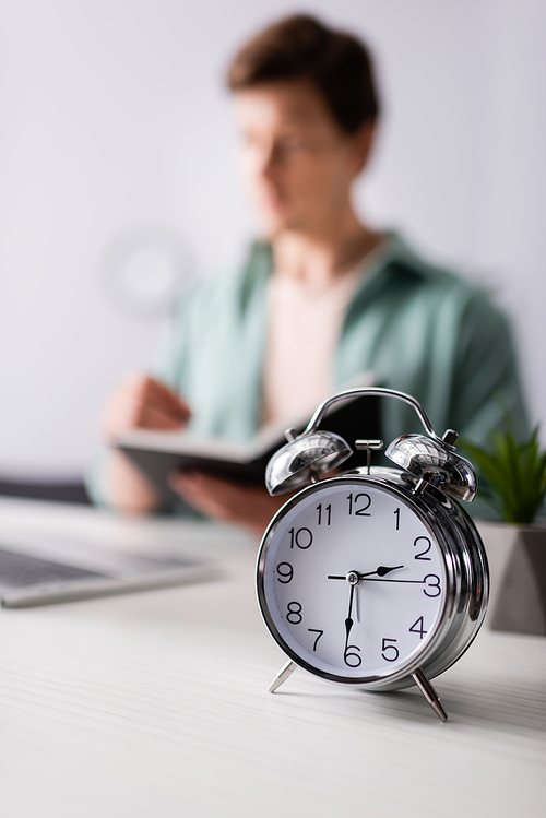 Selective focus of alarm clock on table and man holding notebook near laptop at home, concept of time management