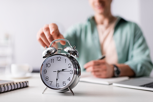 Selective focus of man pulling hand to alarm clock near laptop and notebooks on table, concept of time management