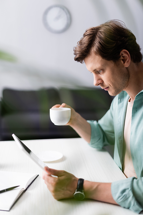 Selective focus of handsome man holding cup of coffee and using digital tablet at table, concept of earning online