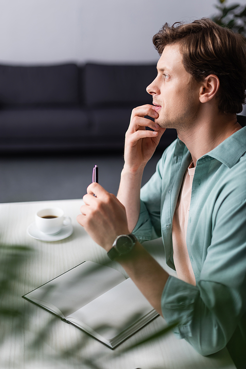 Selective focus of pensive freelancer holding pen near open notebook and cup of coffee at home