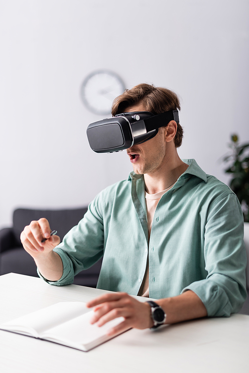 Selective focus of shocked man in vr headset holding pen near notebook on table