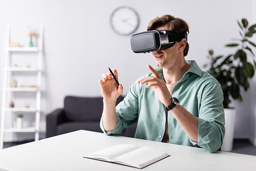 Smiling man in vr headset holding pen near open notebook on table