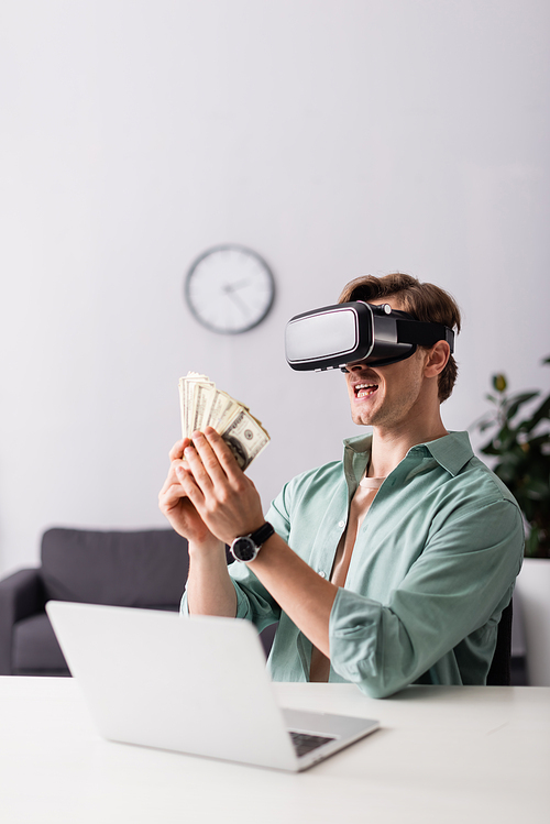 Selective focus of smiling man in vr headset holding cash near laptop on table, concept of earning online