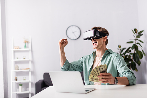 Cheerful man in vr headset showing yeah gesture and holding money near laptop on table, earning online concept