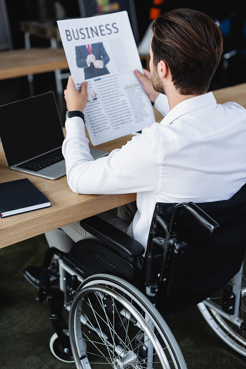 young disabled businessman in wheelchair reading newspaper near laptop with blank screen