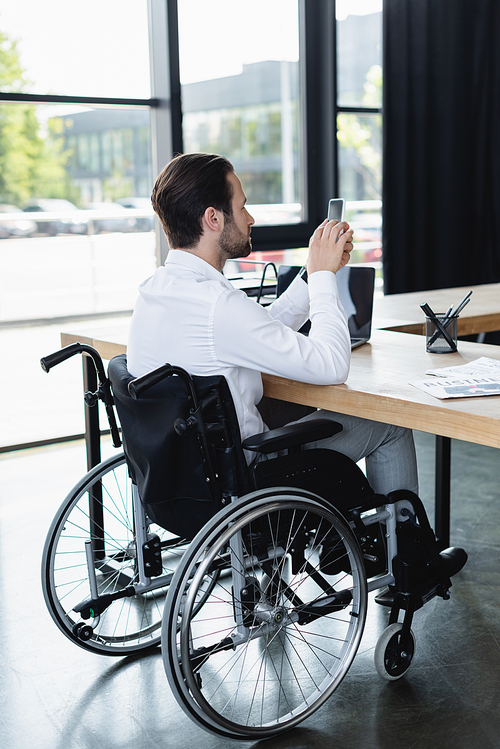 disabled businessman in wheelchair messaging on smartphone near laptop in office