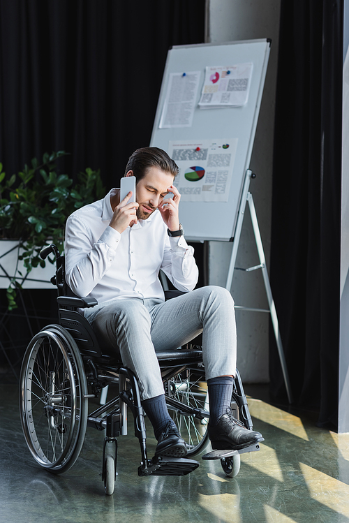 full length view of disabled businessman in wheelchair talking on smartphone near blurred flip chart