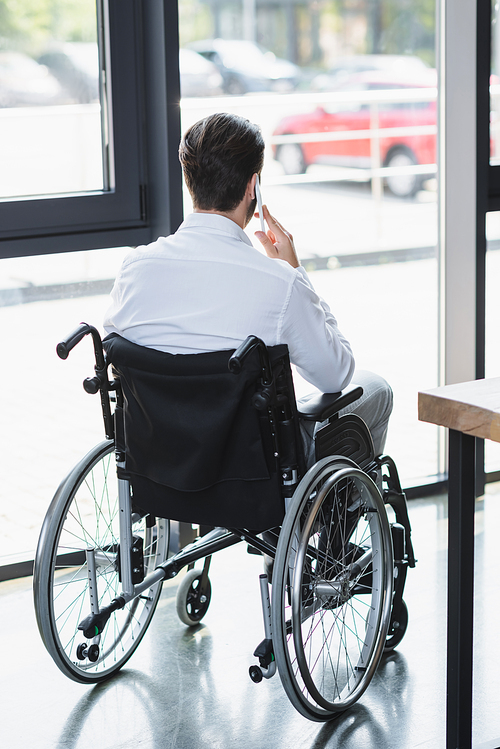 back view of disabled businessman in wheelchair talking on smartphone near window in office