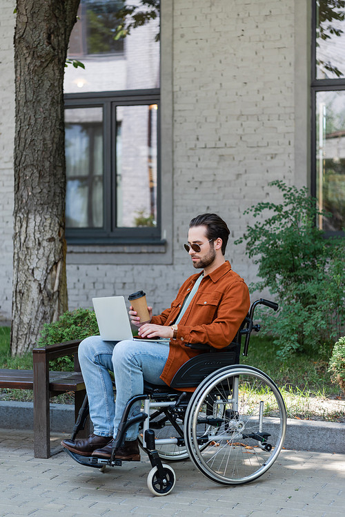 disabled man in sunglasses typing on laptop in wheelchair outdoors