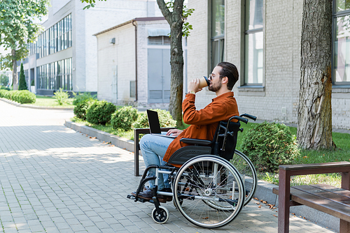 young disabled man sitting in wheelchair with laptop and drinking coffee from paper cup