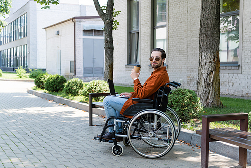 young handicapped man sitting in wheelchair with laptop and takeaway drink outdoors