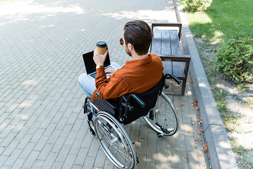 disabled man in wheelchair holding coffee to go while typing on laptop outdoors