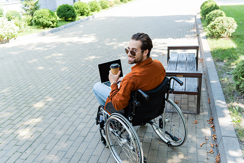 disabled man smiling at camera while sitting in wheelchair with paper cup and laptop