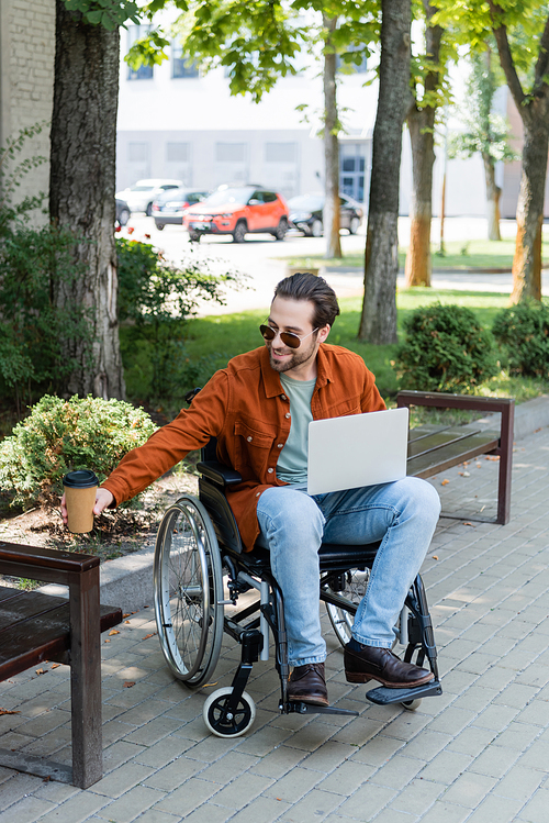 smiling disabled man holding coffee to go while sitting in wheelchair with laptop
