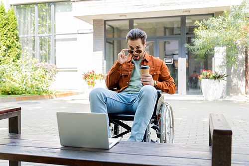 surprised disabled man taking off sunglasses while looking at laptop outdoors