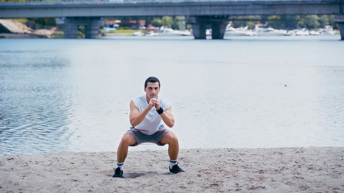 young athletic sportsman doing squat exercise on river shore