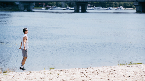 side view of young sportsman jumping with skipping rope on sand near river